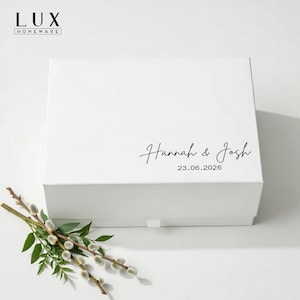 May include: A white gift box featuring the names "Hannah & Josh" and the date "23.06.2026" in black script. The box has a lid and a pull tab. The "LUX HOMEWARE" logo is visible. A sprig of greenery with green leaves and white buds is in the foreground.
