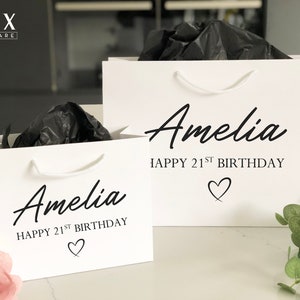 May include: Two white gift bags with black handles and black text. The text on the bags reads "Amelia Happy 21st Birthday" with a heart symbol.
