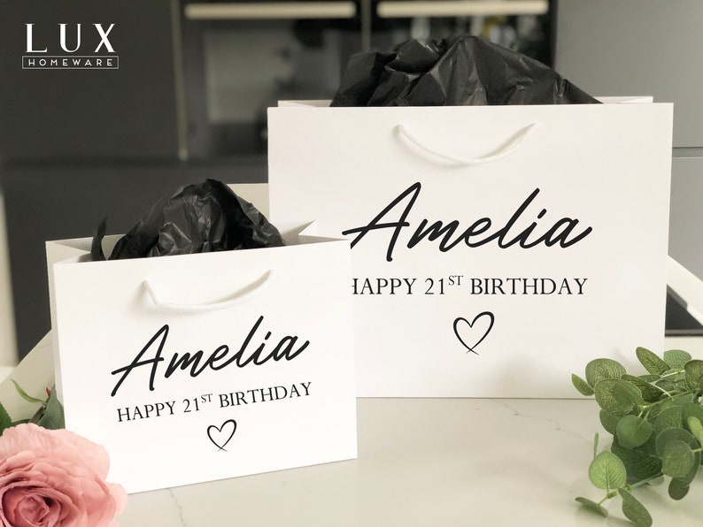May include: Two white gift bags with black handles and black text. The text on the bags reads "Amelia Happy 21st Birthday" with a heart symbol.