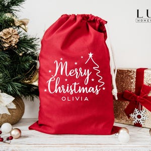 Pode incluir: Uma sacola de presente de Natal vermelha com fecho de cord&atilde;o. A sacola apresenta texto branco que diz "Merry Christmas" e um nome. Uma &aacute;rvore de Natal e um presente embrulhado est&atilde;o no fundo.