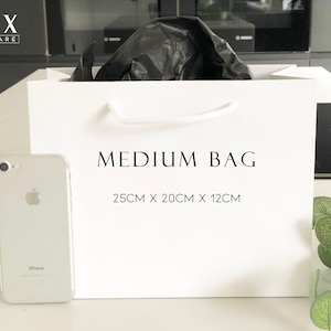 May include: White paper gift bag with black handles and the text "MEDIUM BAG 25CM X 20CM X 12CM" printed on the front. A white iPhone is in the foreground.