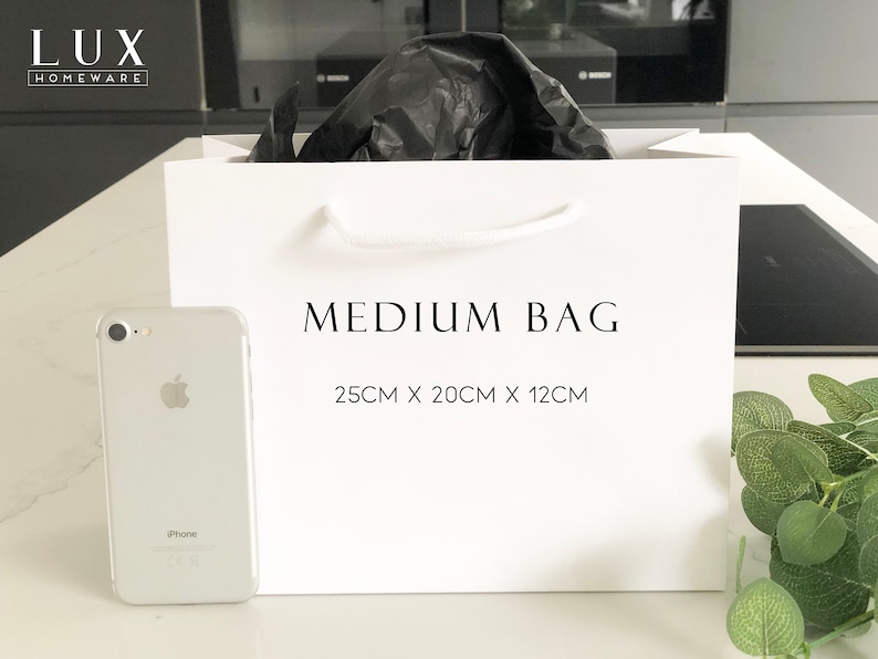 May include: White paper gift bag with black handles and the text "MEDIUM BAG 25CM X 20CM X 12CM" printed on the front. A white iPhone is in the foreground.
