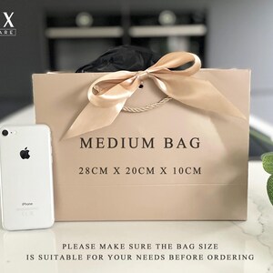 K&ouml;nnte beinhalten: Eine beigefarbene mittelgro&szlig;e Geschenkt&uuml;te mit einer Schleife aus Satinband und Kordelgriffen. Auf der Tasche steht "MEDIUM BAG" und die Ma&szlig;e sind 28 cm x 20 cm x 10 cm. Neben der Tasche liegt ein wei&szlig;es Smartphone.