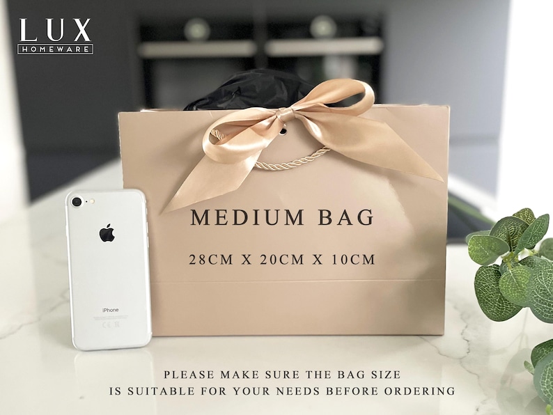 K&ouml;nnte beinhalten: Eine beigefarbene mittelgro&szlig;e Geschenkt&uuml;te mit einer Schleife aus Satinband und Kordelgriffen. Auf der Tasche steht "MEDIUM BAG" und die Ma&szlig;e sind 28 cm x 20 cm x 10 cm. Neben der Tasche liegt ein wei&szlig;es Smartphone.