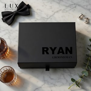 K&ouml;nnte beinhalten: Schwarze Geschenkbox mit dem Namen "RYAN" und dem Wort "GROOMSMAN" auf der Vorderseite. Eine schwarze Fliege, silberne Manschettenkn&ouml;pfe und zwei Gl&auml;ser mit bernsteinfarbener Fl&uuml;ssigkeit sind um die Box herum angeordnet.