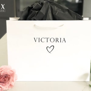May include: White gift bag with a white handle and the text "VICTORIA" with a heart symbol printed on the front. The bag is filled with black tissue paper.