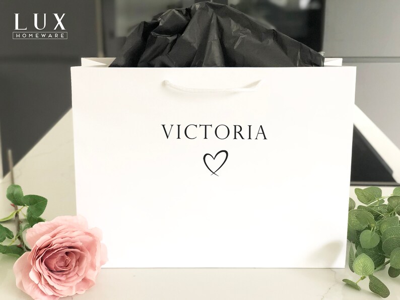 May include: White gift bag with a white handle and the text "VICTORIA" with a heart symbol printed on the front. The bag is filled with black tissue paper.