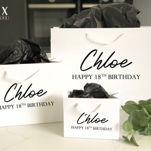 May include: Three white gift bags with black handles and the text "Chloe Happy 18th Birthday" printed on them. The bags are filled with black tissue paper and are sitting on a white surface.