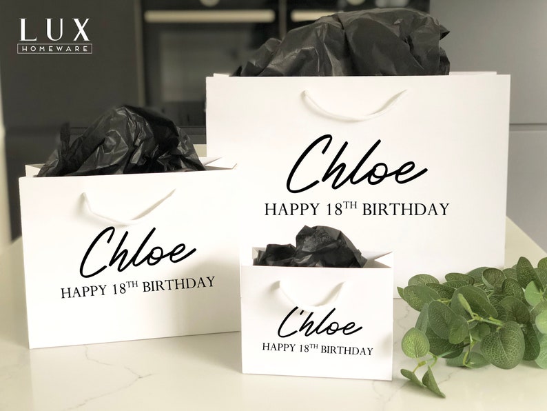 May include: Three white gift bags with black handles and the text "Chloe Happy 18th Birthday" printed on them. The bags are filled with black tissue paper and are sitting on a white surface.