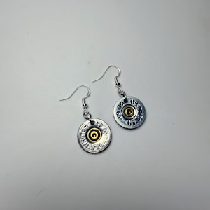 May include: A pair of handmade earrings made from silver-coloured bullet casings with gold-coloured centres. The earrings have a hook closure and are stamped with the word "REMINGTON".