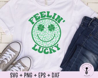 May include: A white t-shirt with a green shamrock design that says "Feelin' Lucky" in a retro font. The shamrock is a smiley face with a leopard print pattern.