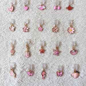 May include: A collection of pink and gold charms in various shapes, including hearts, flowers, and a seahorse. Each charm is attached to a clear plastic clip. The charms are displayed on a white, textured surface.