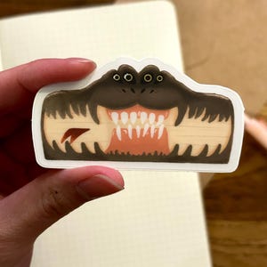 May include: A sticker featuring a cartoon alligator mouth with sharp white teeth, brown gums, and dark brown skin. The sticker is held against a notebook with a grid pattern.