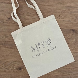 Könnte beinhalten: Eine beige Canvas-Tragetasche mit schwarzen Henkeln und einem schwarzen grafischen Druck von stilisierten Blumen und dem Text "DIFFERENT is beautiful".