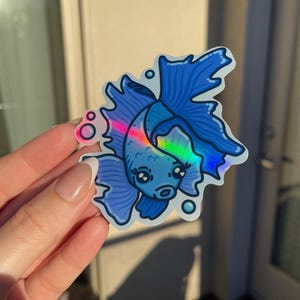 May include: A blue iridescent sticker of a cartoon fish with large eyes and a single fin. The fish is surrounded by bubbles.