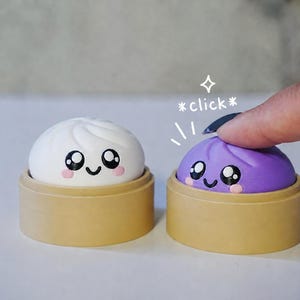 May include: Two small, round, smiling dumpling-shaped toys, one white and one purple, each resting in a tan circular base. The purple toy is being pressed down, with the text *click* and a small star icon visible above it.