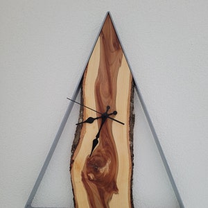 Könnte beinhalten: Eine dreieckige Wanduhr mit einem Zifferblatt aus Naturholz und einem grauen Metallrahmen. Die Uhrzeiger sind schwarz. Die Holzmaserung ist sichtbar, mit Schattierungen von hellbraun und rötlich-braun. Die Uhr ist an einer weißen Wand befestigt.