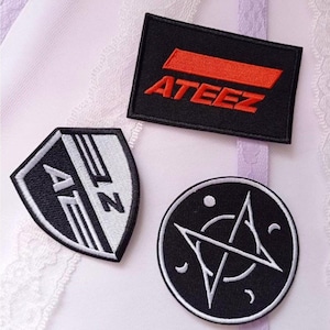 May include: Three embroidered patches with different designs. One is a black rectangle with the word "ATEEZ" in red. Another is a shield shape with black and white stripes and the letters "A" and "Z". The third is a black circle with a white star design.