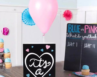 May include: A black chalkboard sign with a white heart that says "it's a" surrounded by blue and pink hearts. A pink balloon is attached to the top of the sign. The sign is on a table with a chalkboard sign that says "Blue or Pink? What do you think?" with tally marks on the right side.