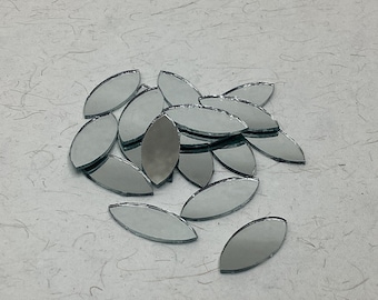 May include: A pile of small, silver, oval-shaped mirror tiles. The tiles are arranged in a loose pile on a white, textured surface.