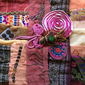 May include: A patchwork quilt made of various fabrics in shades of pink, brown, and beige. The quilt features a variety of textures, including lace, embroidery, and sequins. A pink and white embroidered swirl, a brown fish with a green jewel, and a pink crocheted flower are visible in the center of the quilt.