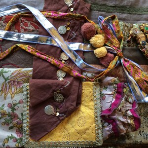May include: A close-up of a fabric patchwork with various textures and colors, including brown, yellow, pink, and blue. The fabric is decorated with gold coins, sequins, and pom-poms. A small bird-shaped ornament with turquoise and gold accents is also visible.