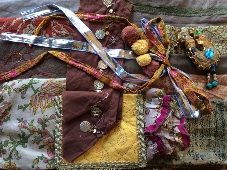 May include: A close-up of a fabric patchwork with various textures and colors, including brown, yellow, pink, and blue. The fabric is decorated with gold coins, sequins, and pom-poms. A small bird-shaped ornament with turquoise and gold accents is also visible.