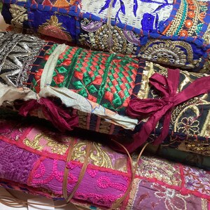 May include: A stack of colorful fabric rolls with intricate embroidery and embellishments. The rolls are tied together with a burgundy ribbon and feature a variety of patterns and textures.