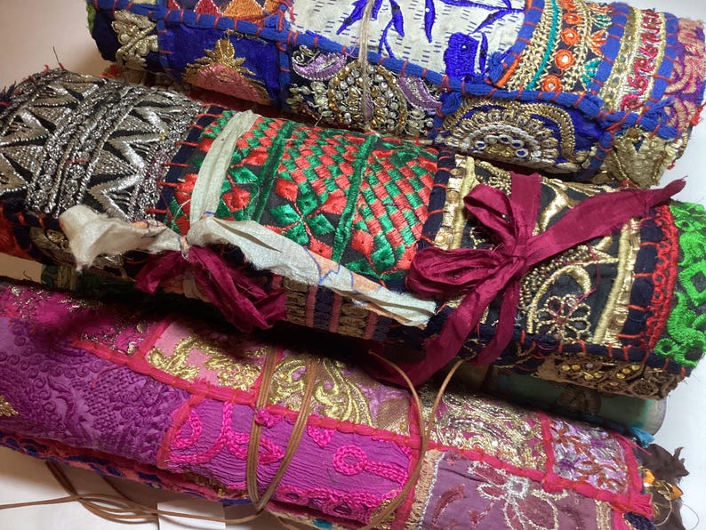 May include: A stack of colorful fabric rolls with intricate embroidery and embellishments. The rolls are tied together with a burgundy ribbon and feature a variety of patterns and textures.