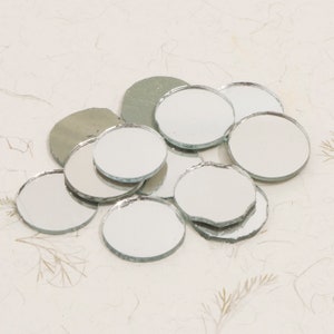 May include: A pile of round, silver-coloured mirror tiles. The tiles are various sizes and are scattered on a white surface.