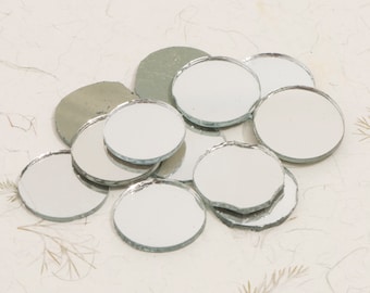 May include: A pile of round, silver-colored mirror tiles. The tiles are various sizes and are scattered on a white surface.