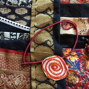 May include: A close-up of a red, white, and orange fabric flower with a spiral design. The flower is attached to a red cord and is lying on top of a colorful fabric patchwork.