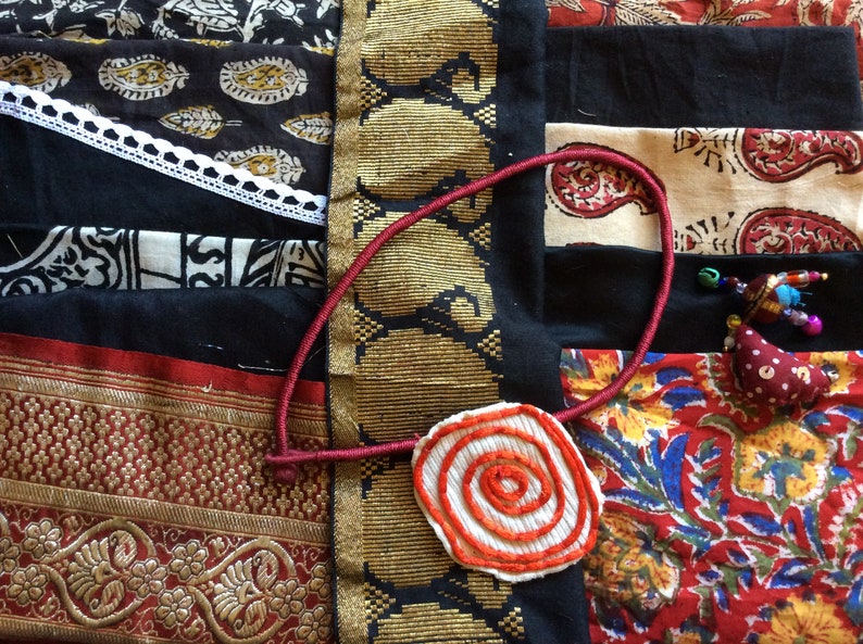 May include: A close-up of a red, white, and orange fabric flower with a spiral design. The flower is attached to a red cord and is lying on top of a colorful fabric patchwork.