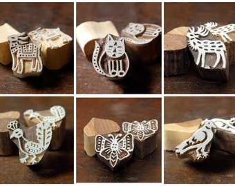 May include: A collection of six wooden block stamps with animal designs. The stamps feature a variety of animals, including an elephant, cat, deer, peacock, butterfly, and bird. Each stamp is carved with intricate details and painted white.