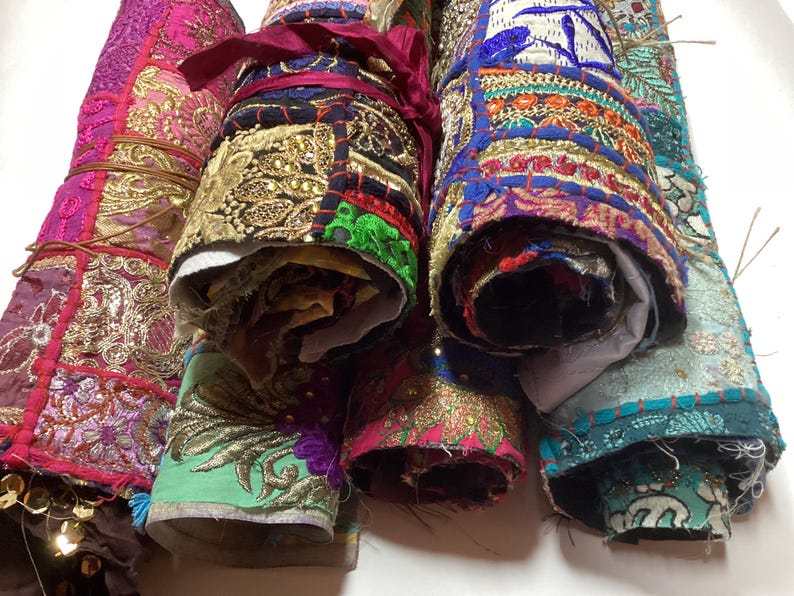 May include: Five colorful fabric rolls with intricate embroidery and embellishments. The rolls are made of different fabrics, including velvet, silk, and cotton. The fabrics are all in various shades of pink, blue, green, and gold.