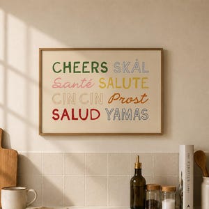 Puede incluir: Un cuadro rectangular con marco de madera fina que muestra las palabras CHEERS, SKÅL, Santé, SALUTE, CIN CIN, Prost, SALUD y YAMAS en varios colores y fuentes, dispuestas en cuatro filas sobre un fondo de color crema claro.