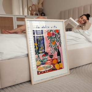 May include: A framed art print with the words "la dolce vita" features a colourful still life. The artwork depicts a table with fruit, wine, and flowers. The frame is light-coloured with a dotted border. The print is displayed in a bedroom.
