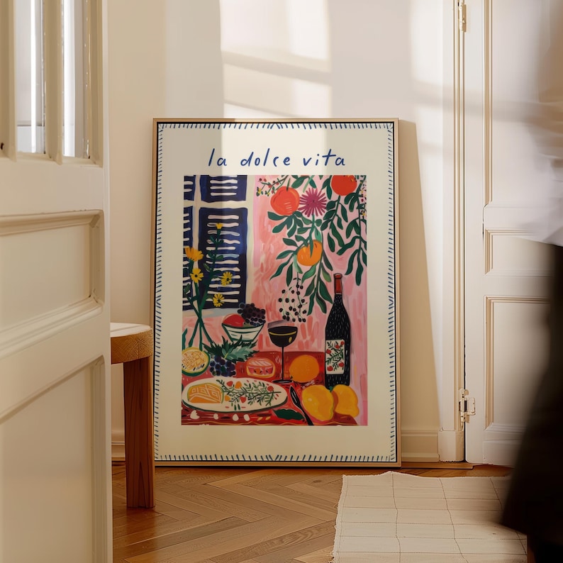 May include: A framed art print with the words "la dolce vita" in blue script. The colourful illustration depicts a table setting with a bottle of wine, fruit, and glasses. The background features a pink wall and a window with blue shutters.