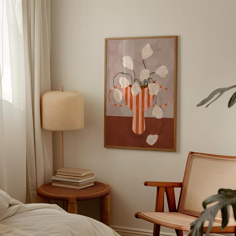 May include: A framed art print featuring a vase with white flowers and a striped design in shades of orange and red. The artwork has a neutral background and is displayed in a room with a lamp and a side table.