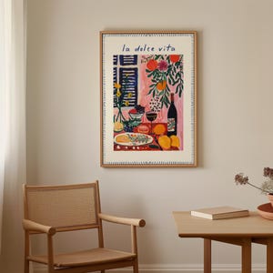 May include: A framed art print titled "la dolce vita" shows a still life with a bottle of wine, fruit, and a plate of food. The artwork uses a vibrant colour palette with pink, orange, and green hues, creating a cheerful and inviting atmosphere.