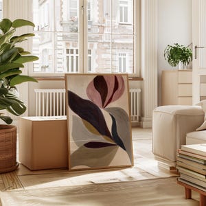 Peut inclure: Une impression d'art floral abstrait encadrée. L'œuvre présente une grande fleur stylisée dans des tons de rose, de violet et de bleu sur un fond crème. Le cadre est de couleur bois clair. L'impression est appuyée contre un cube aux tons neutres.