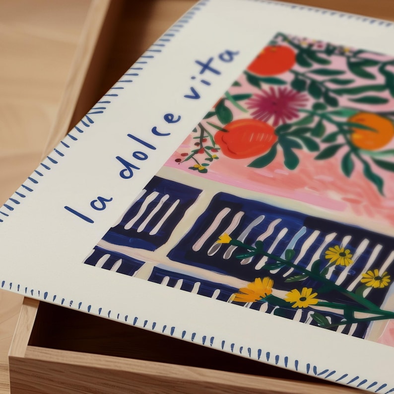 May include: An art print with the words "la dolce vita." The illustration shows oranges, flowers, and a blue and white striped balcony. The print has a white border with a dotted blue pattern. The artwork is in a wooden frame.