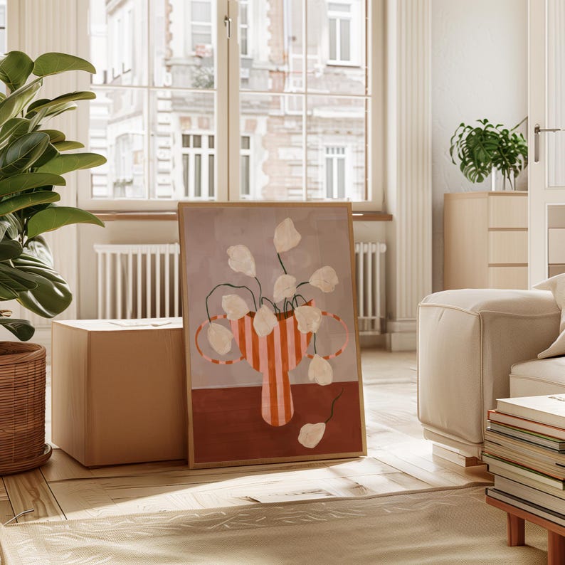 May include: A framed art print featuring a still life of white flowers in a striped vase. The vase is orange and white, set against a gray background with a brown base. The artwork is displayed in a bright, airy room with a large window.