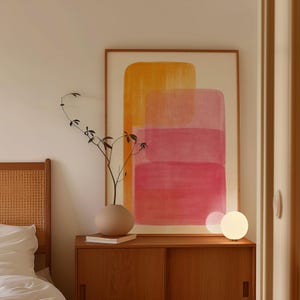 May include: A wooden framed abstract art print with orange and pink rectangular shapes hangs above a wooden cabinet. A beige vase with a sprig of greenery and a small, round, white lamp sit on the cabinet. A bed frame is visible on the left.