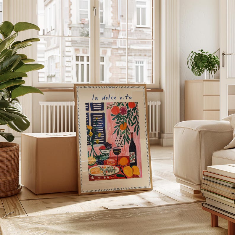 May include: A framed art print with the words "la dolce vita" depicts a vibrant illustration of a table setting with food, wine, and fruit. The artwork has a pink background and a wooden frame. The print is displayed in a bright room with a large window.