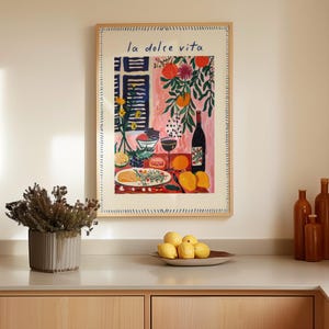 May include: A framed art print with the text "la dolce vita" in blue script. The artwork depicts a still life with a wine bottle, fruit, and a glass of wine. The frame is a light wood colour.