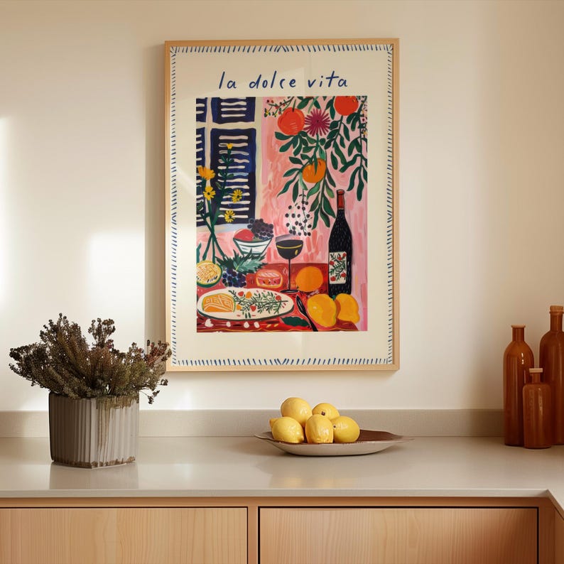 May include: A framed art print with the text "la dolce vita" in blue script. The artwork depicts a still life with a wine bottle, fruit, and a glass of wine. The frame is a light wood colour.