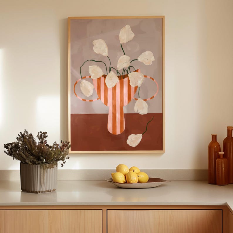May include: A framed art print featuring a vase with white flowers and a striped design in shades of orange and pink. The artwork is displayed above a countertop with a decorative plant, a bowl of lemons, and brown bottles.