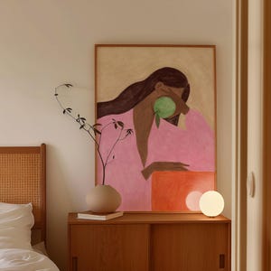 Op de afbeelding: Een ingelijste kunstdruk toont een persoon in een roze gewaad die een groene vrucht vasthoudt. Een houten kast met een beige vaas en een kleine witte lamp staat voor het kunstwerk. Een takje groen staat in de vaas.