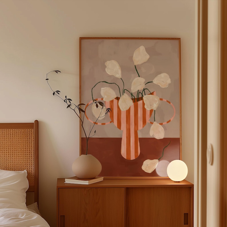 May include: A framed abstract art print featuring a vase with vertical orange and pink stripes, holding white flower-like shapes. A small, round, beige vase and a spherical lamp sit on a wooden cabinet. A sprig of greenery is in the foreground.